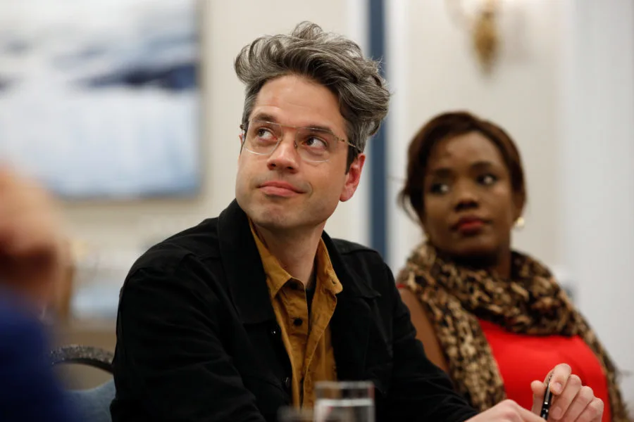 A man with a light warm golden skin tone looks up. He has grey and brown-streaked hair and is wearing glasses and a black jean jacket with a light brown button up shirt. A Black woman with a deep skin tone sits next to him. 