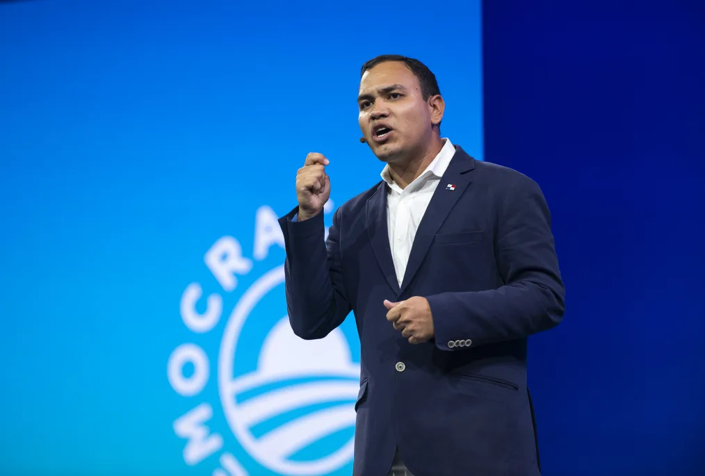 A middle aged man with medium-deep skin who has fairly short slick black hair. He has a navy suit on, with a white button up underneath. He has a mic coming behind his right ear and is giving a speech. In the background is a light blue and dark blue gradient with a Obama Foundation logo off to the left side.