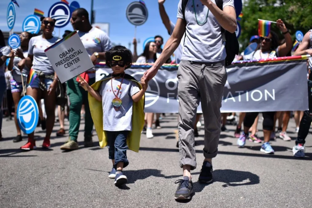 
A young kid with a light-medium skin tone wears a batman mask and holds a poster that reads "Obama Presidential Center" while holding someone's hand with a light skin tone in the pride parade 