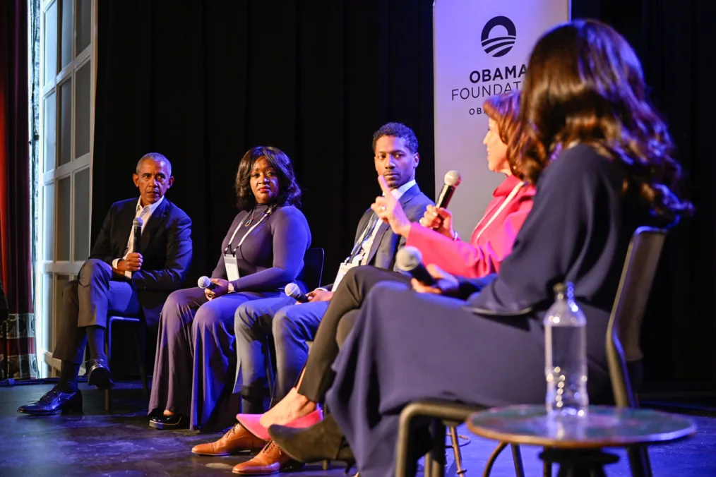 President Obama sits on stage holding a microphone and looking right at four people of varying skin tones and clothing, also seated.