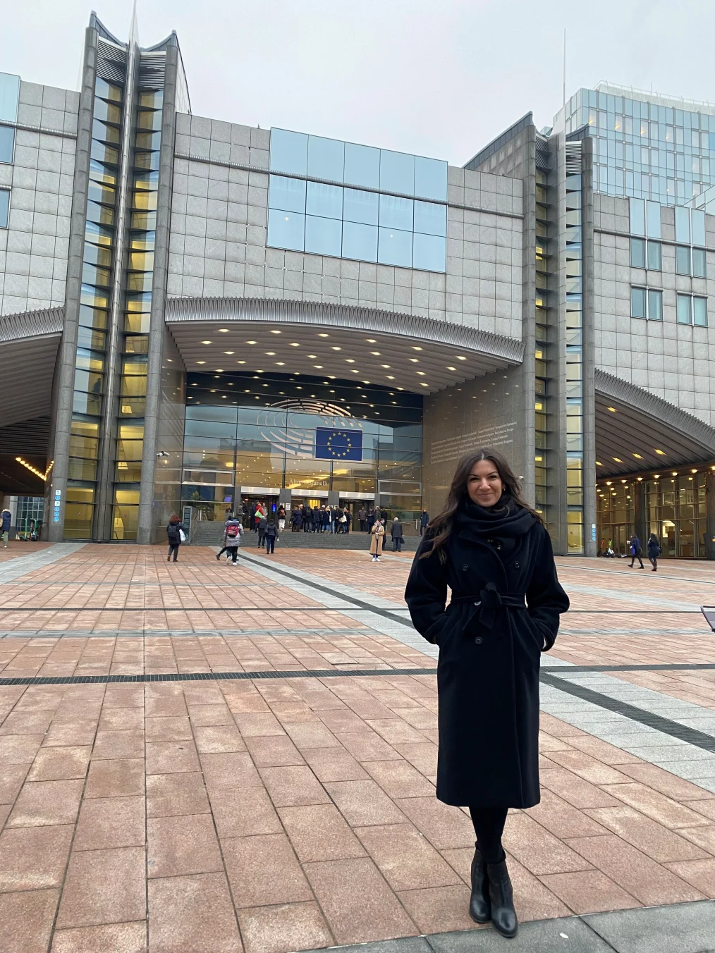 Ornella Cosomati, holds a closed lip smile as she stands outside the European Parliament building. In the background, people are walking in. There is a flag of Europe in the center of the building.