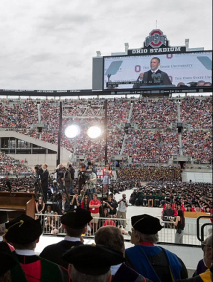 President Obama is dressed in a black robe,and is being shown on a stadium screen at Ohio State University's commencement ceremony. The stadium seats below the screen are filled with people, there are two bright lights in the center of the photo and several men wearing ceremonial tams are shown from behind. 