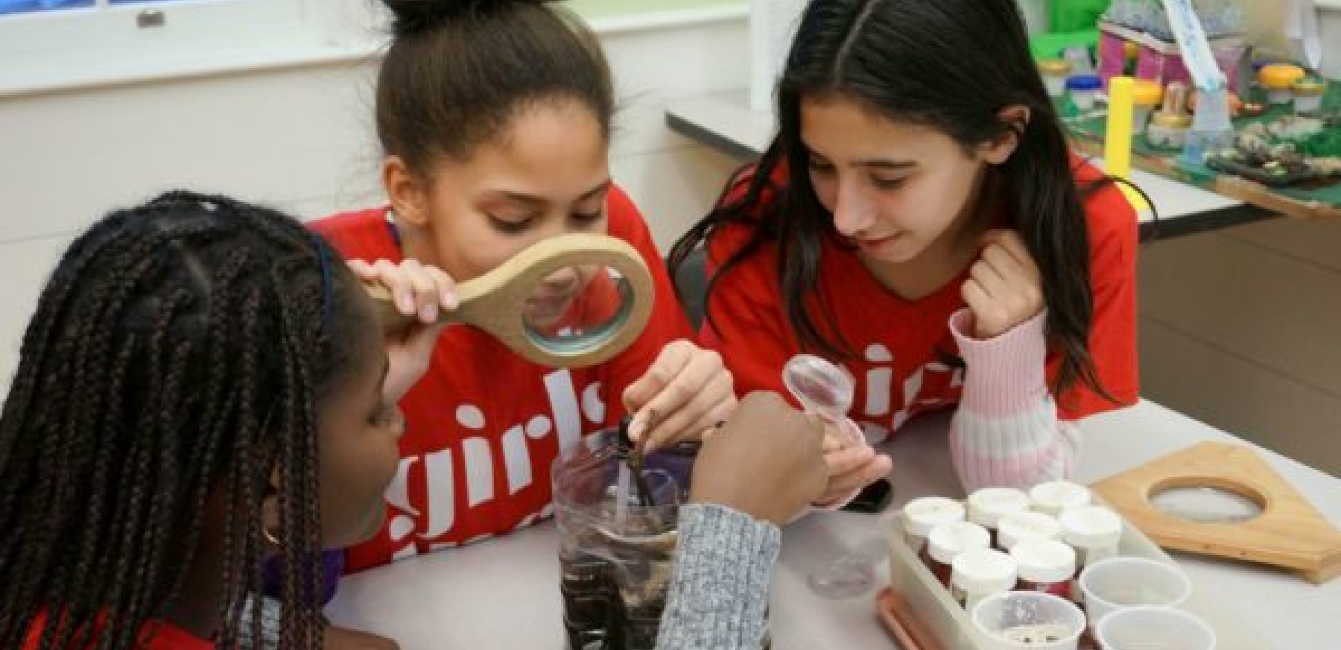 In this picture, three young girls with different skin tones are shown with 
magnifying glasses are looking into a cut-in-half bottle of brown water.