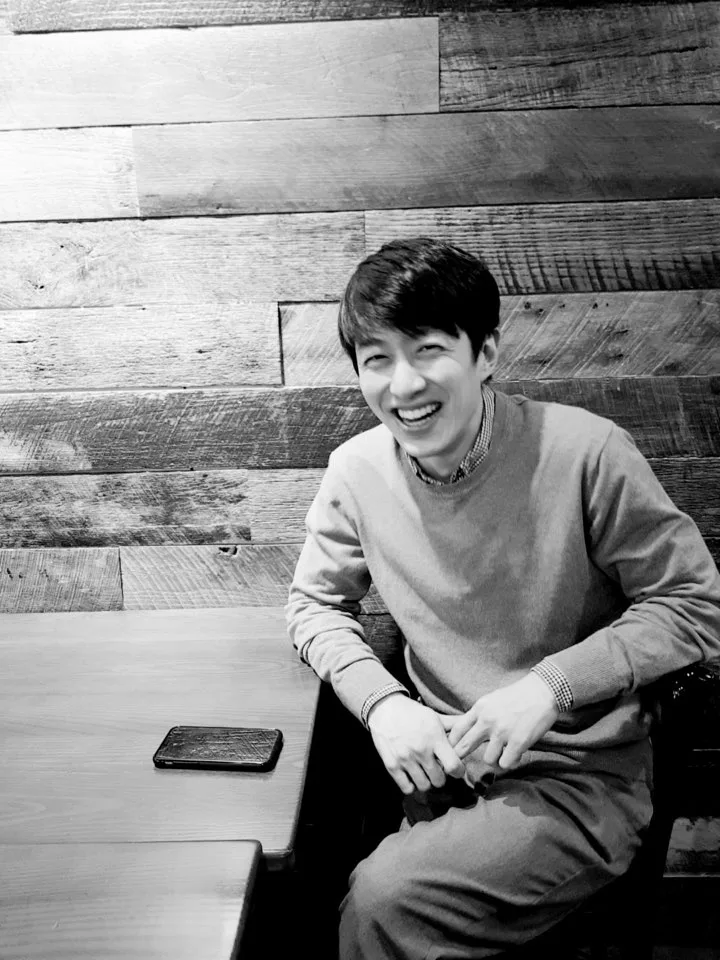 In a black and white photo, Joseph Lim, an Asian man, smiles as he sits at a table.
