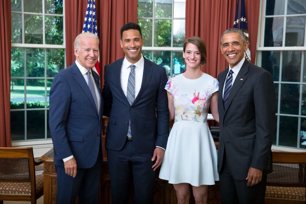 Then Vice President Joe Biden, Devin Hampton, Katy Mason, and President Obama pose for a photo in the Oval Office. All men are wearing navy suits and a variation of blue-striped ties and are a range of skin tones. Katy is wearing a white watercolor dress and has short brown hair and a light skin tone.