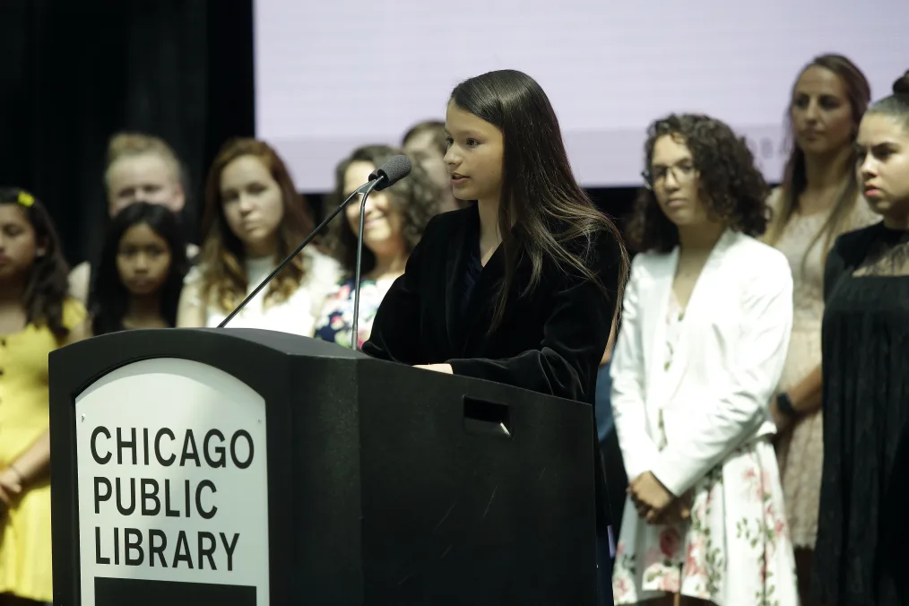 

A young girl with a light skin tone with black hair stands in front of a podium that reads "Chicago Public Library" in front of a group of young girls and women ranging from deep medium to light skin tones
