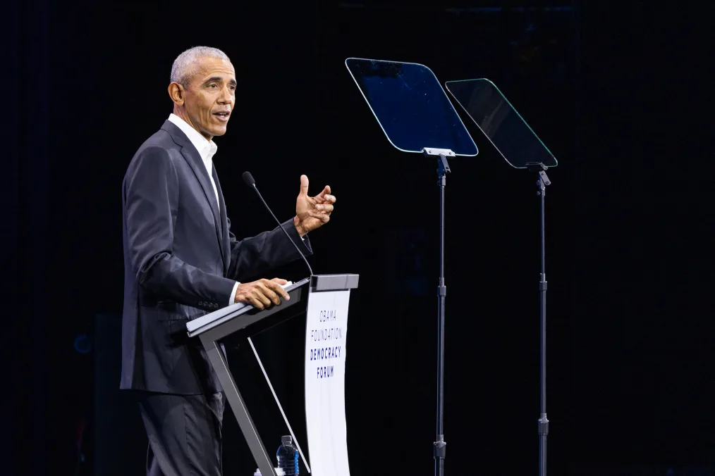 President Obama stands behind a podium and speaks to an audience. Two clear teleprompters are in front of him. A screen behind him reads, “Obama Foundation Democracy Forum.” He is wearing a black suit without a tie. 