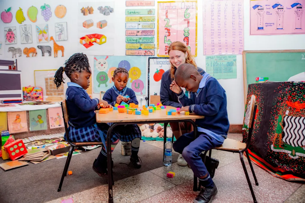 A woman with shoulder length red hair sits at a child’s table with three little ones with deep skin tones. They are wearing navy school uniforms and play with blocks on a table. Art and posters are in the background.