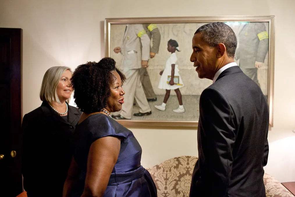 President Obama stands talking to an older woman in a blue dress with a deep skin tone and brown curly hair in front of a painting of Ruby Bridges, a young girl with a deep skin tone. Behind them is an older woman with a light skin tone and straight, short gray hair in a black dress