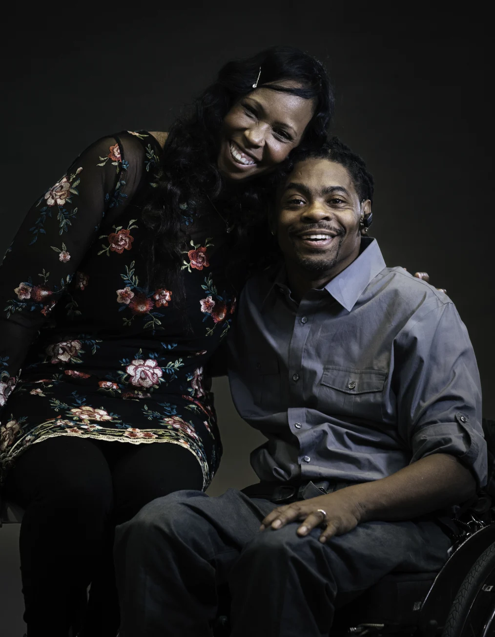 This picture shows a man and a woman with deep skin tones smiling toward the camera. The man
is sitting in a wheelchair with an earpiece attached to his ear while the woman leans in and hugs him.