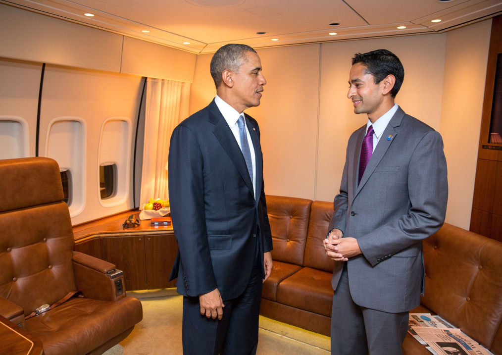 Two men stand at the center of the image facing each other: one is President Barack Obama, the other is Anuj Gupta. President Obama is wearing a dark blue suit with a white collared shirt, and a light blue tie. He has an American flag pin fastened to the left lapel of his suit. He is in mid-conversation. Anuj is a brown-skinned man with short dark black hair. He is wearing a gray suit with a white collared shirt, and a purple tie. He has an Obama campaign pin fastened to the left lapel of his suit. Anuj is clutching his hands together and smiling at President Obama. In the image, the two men are located on Air Force One. On the plane are brown leather reclining chairs and a brown wooden desk. On one of the chairs are three newspapers – publication and copy indiscernible. The walls of the plane are beige. There are three plane windows, each of which are fully open. 