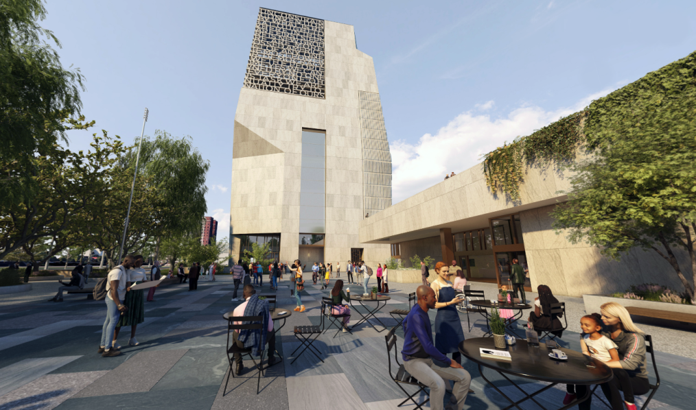 The image is a rendering of the John Lewis Plaza. The Plaza is outdoors and the ground is covered in gray concrete pavers. There are trees on the left hand side of the plaza. In the background of the plaza is the Obama Presidential Center Museum. On the right hand side of the image is the entrance to the Obama Presidential Center Forum.  On the pavers of the plaza are tables and chairs. Across the Plaza are people of different races and ethnicities, standing, sitting, and walking on the Plaza.