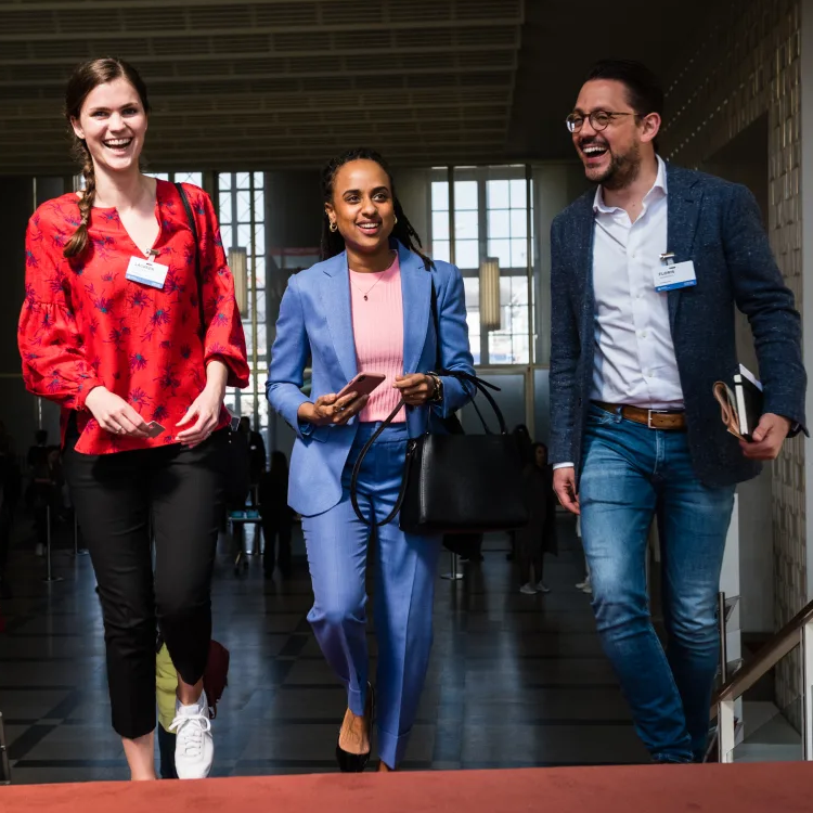 A woman with a light skin tone walks up the stairs with another woman with a medium skin tone and by her side is a man with a light-medium skin tone and glasses. They all smile while wearing business attire