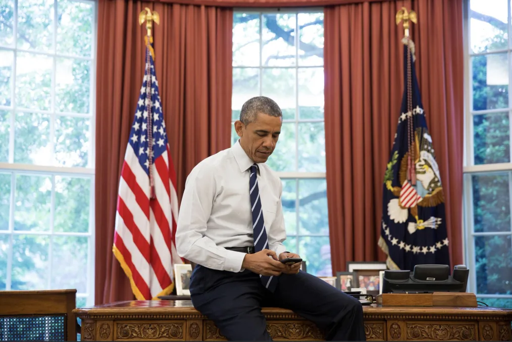 Barack Obama sits on a wood desk in the Oval Office typing on his cell phone. Red curtains and flags representing the U.S. are behind him in front of large windows.