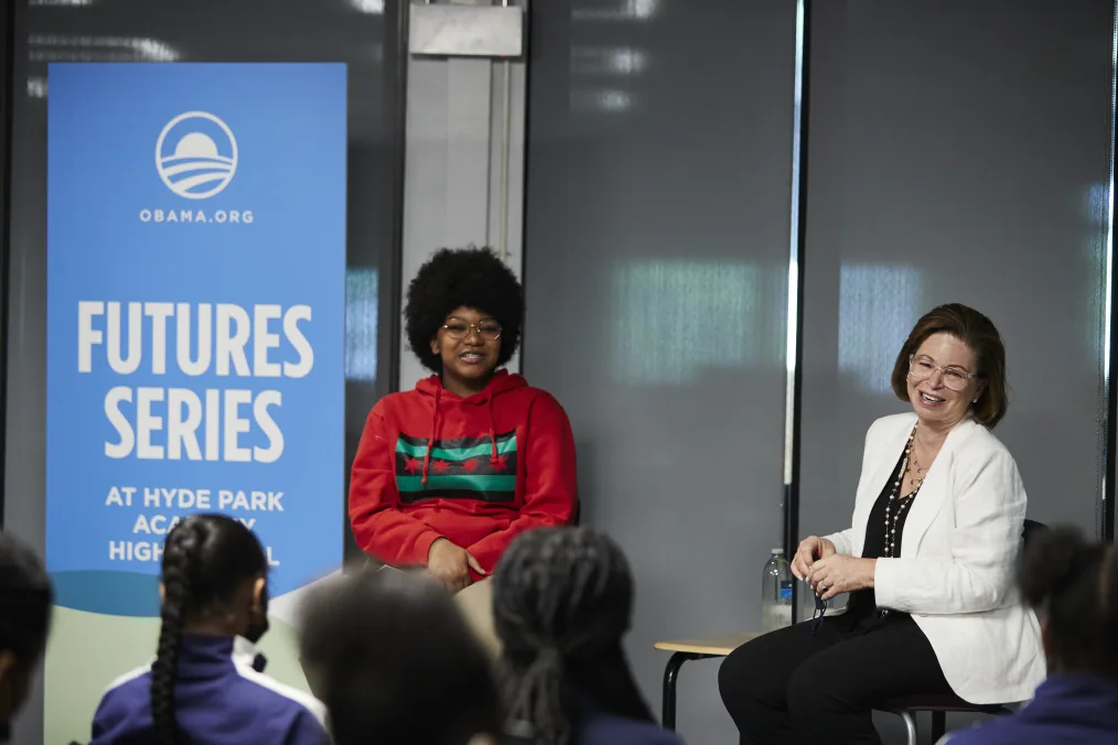 Valerie Jarrett sits smiling and speaking to a crowd of young students with dark hair. Sitting beside her, a young person with a medium deep skin tone who wears a red sweatshirt that displays a red, green and black Chicago flag. Next to them, a banner that reads 'FUTURES SERIES AT HYDE PARK ACADEMY HIGH SCHOOL." 