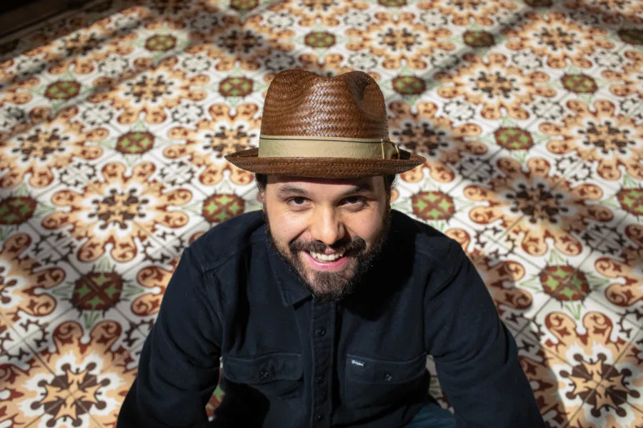 A portrait shows a man with a light medium skin tone sit on a floor with an abstract rug that is green, white, yellow, brown, and burgandy. He is smiling and wearing a brown fedora and a black button up jacket. 