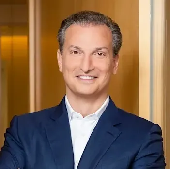 A white man with gray hair wearing a blue suit. He smiles for the camera. His arms are crossed. There is a golden color to the background.