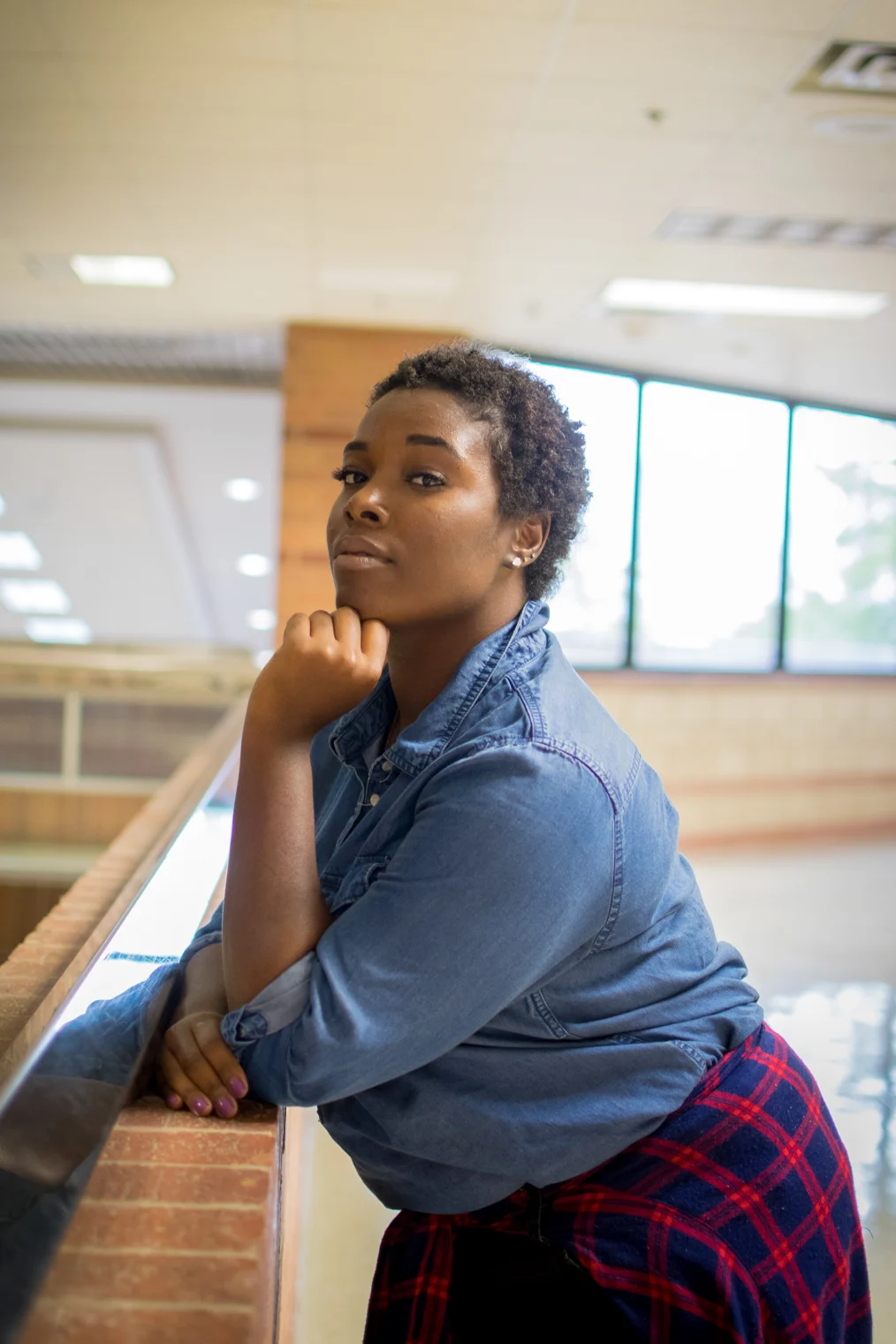 A deep skin toned Black woman with short cut hair is leaning on a short wall with her body turned to the left. She is facing the camera with a straight expression and her left hand in a loose fist under her chin. She is wearing a denim button up shirt with a blue and red flannel tied around her waist, pink nail polish and several earrings in her left ear. The background appears to be a hallway within a building. 
