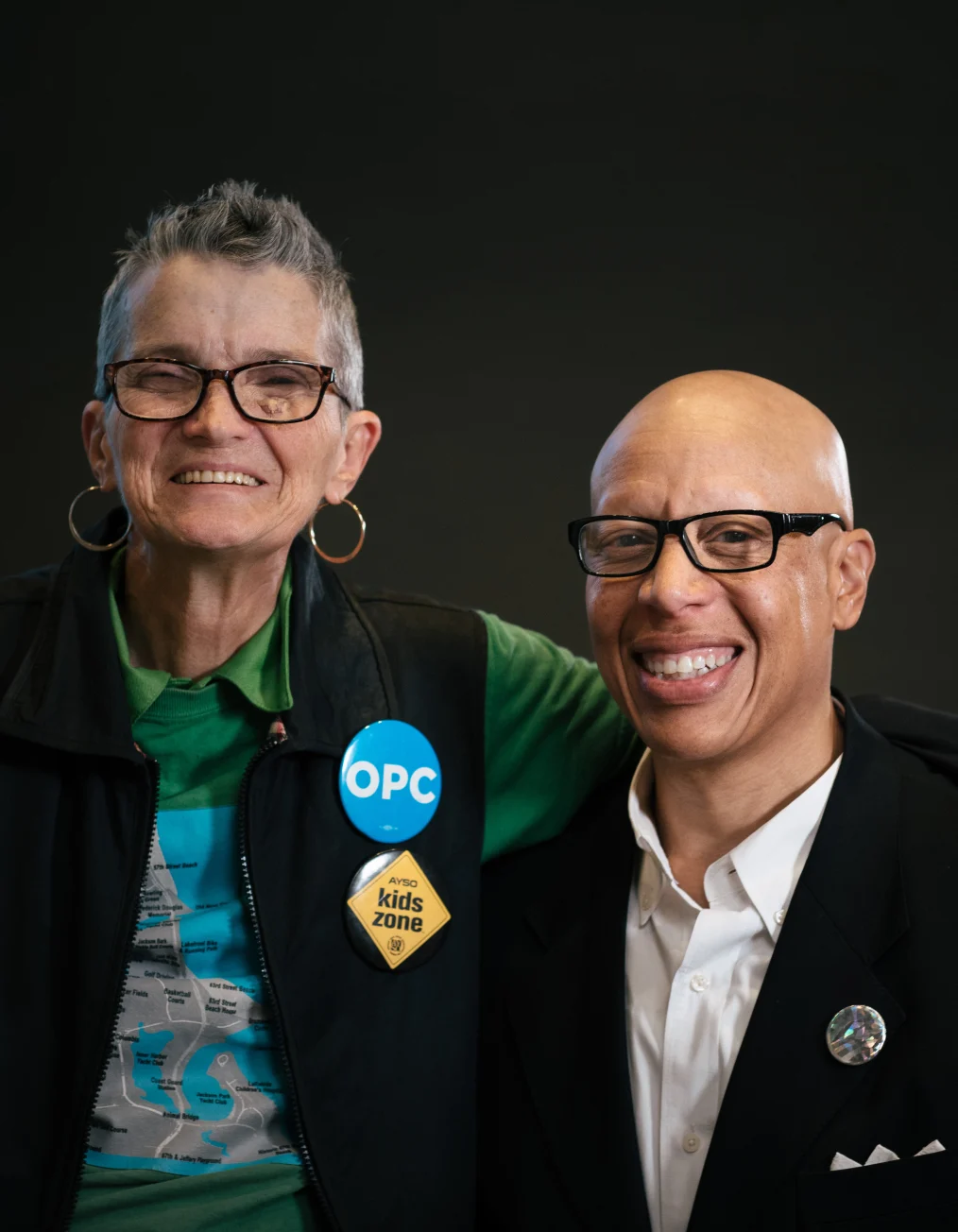 In this picture, a man and woman with different skin tones pose smiling in front of a black backsplash 
 facing forward at the camera. The man has a medium skin tone, while the woman has a light skin tone.