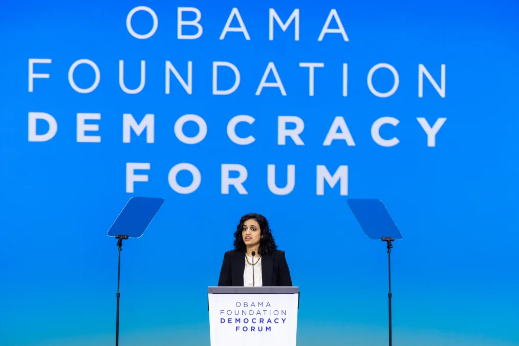 Aleena Agarwal, an indian woman with a medium skin tone and short black hair stands behind a podium with two clear teleprompters in front of her. The front of the podium reads, “Obama Foundation Democracy Forum.” A screen in the background says the same. 