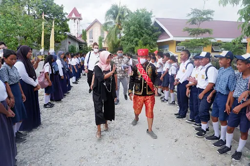 Ayu Kartika Dewi smiles and waves as she walks between two lines of more than 20 Indonesian students. All are a range of medium deep skin tones. The students are wearing uniforms and Ayu is wearing a black garb and pink hijab.