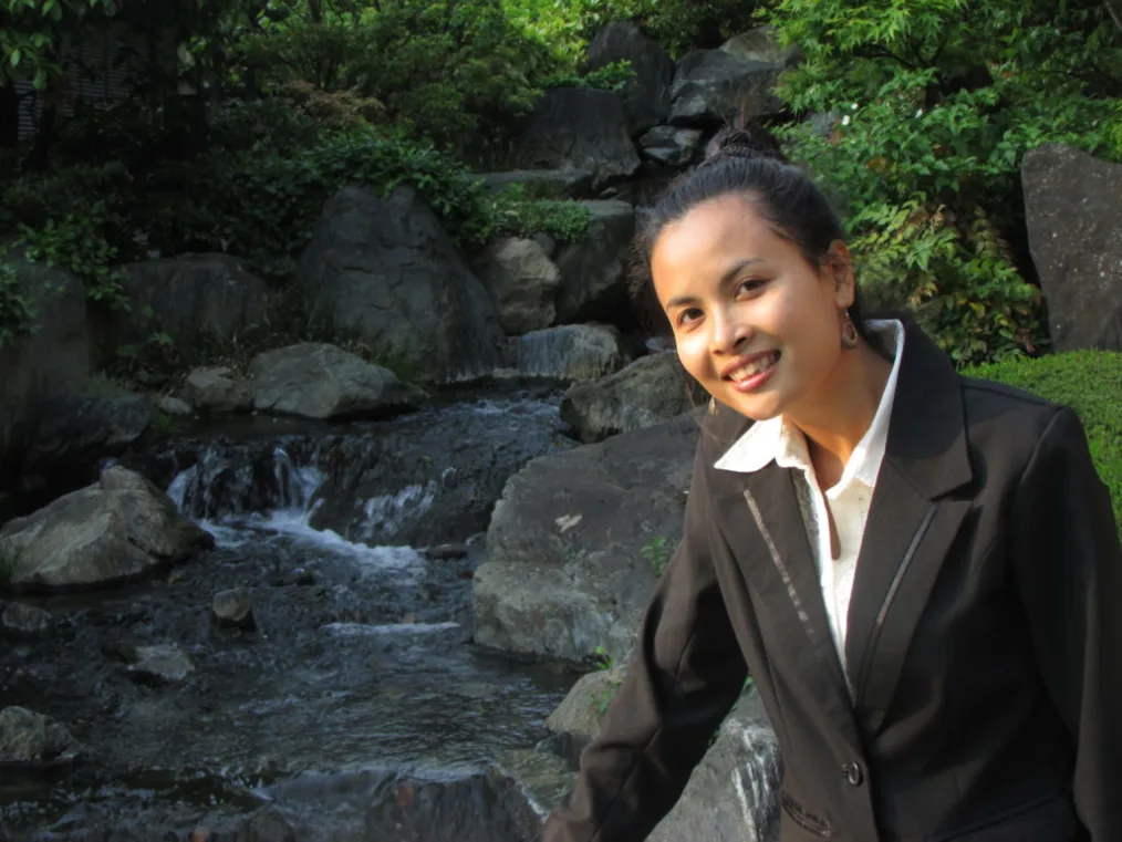 A woman with a light medium skin tone stands in front of a stream of water with rocks and leaves around it. She is wearing a ponytail, a black suit jacket, and a loose white button up.