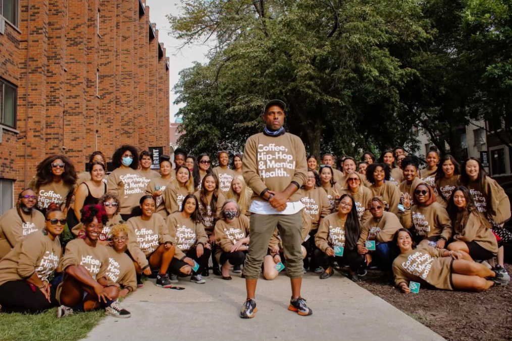 Christopher LeMark, a Black man with a medium skin tone stands in front of a large group of people as they pose for a picture. All have a range of light to deep skin tones. Each person is wearing a brown crew neck that reads, “Coffee, Hip-Hop, & Mental Health.”