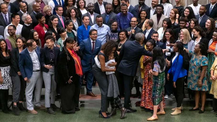 In the center of the photo, Michelle Obama is wearing a gray jumpsuit with blue heels and embracing a smiling medium brown skin toned child who is wearing a black and shite checkered dress, gray and black horizontal striped tights and brown ankle boots. To the right of them, Barack Obama is turned away from the camera wearing a dark colored suit, shaking hands with a woman standing on risers. Michelle and Barack are surrounded by about 50 smiling people of various skin tones who are standing on 5 levels of risers. 