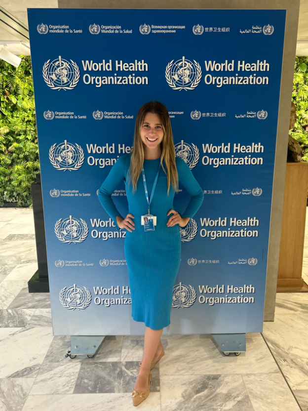 Liza Zaruba, a woman with a light skin tone and long blonde hair, stands in front of a blue and white gradient backdrop that reads, “World Health Organization.” She is wearing a long sleeve blue dress. 