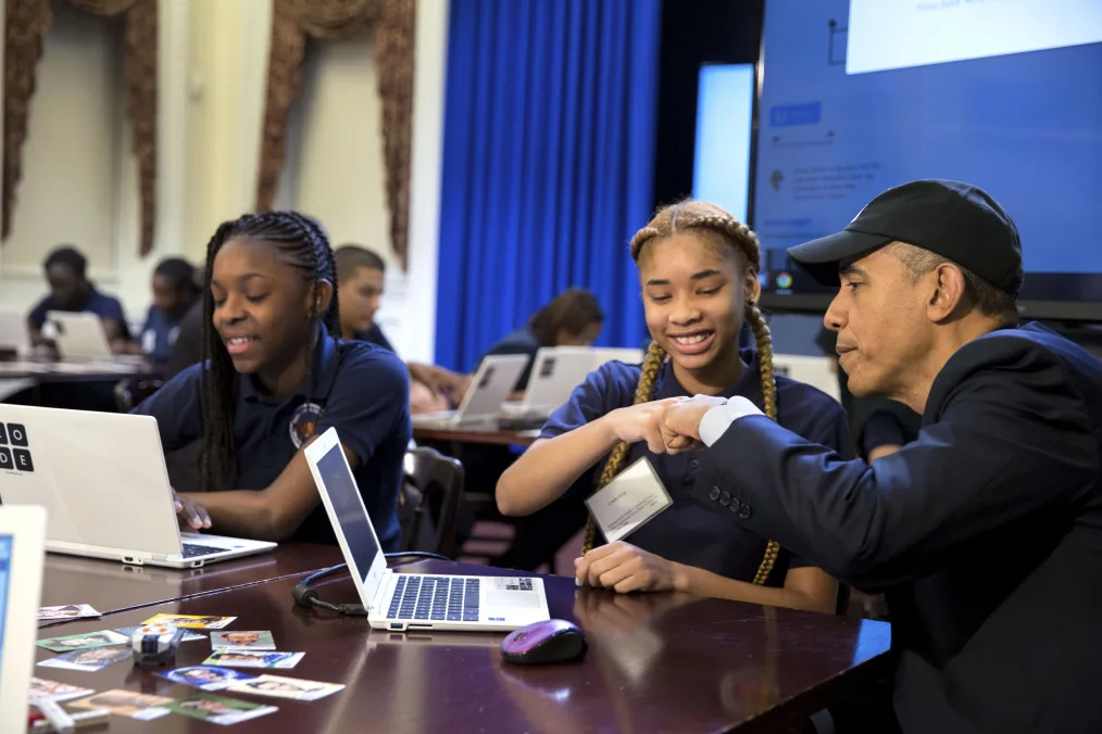 President Obama fist bumps a young girl with a medium skin tone while sitting in front of a computer. He is sitting at a table with another young woman with a deep skin tone. 