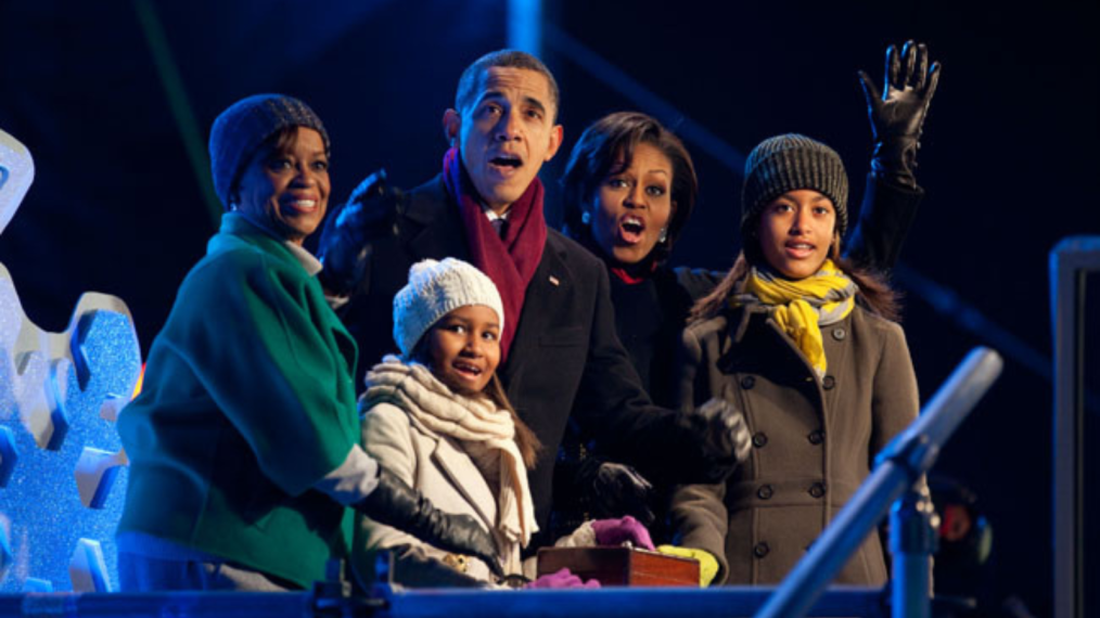 An image of the Obama Family waving to a crowd outside during a winter event. They are all wearing coats and scarves. The family includes: Marian Robinson, Sasha Obama, Barack Obama, Michelle Obama, and Malia Obama