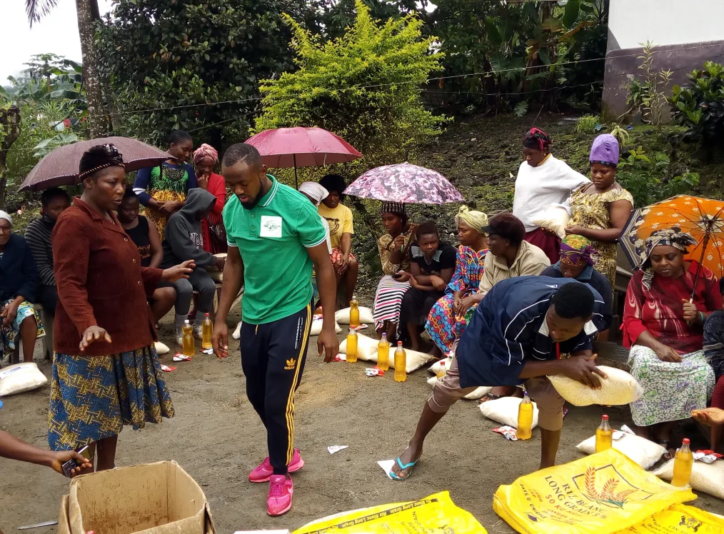 Obama Leader Njeke Joshua Egbe, a man with a dark skin tone and low black hair, teaches an organic farming course to people in his community. He is surrounded by a group of people with a range of ages. They have dark skin tones and are holding umbrellas outdoors.