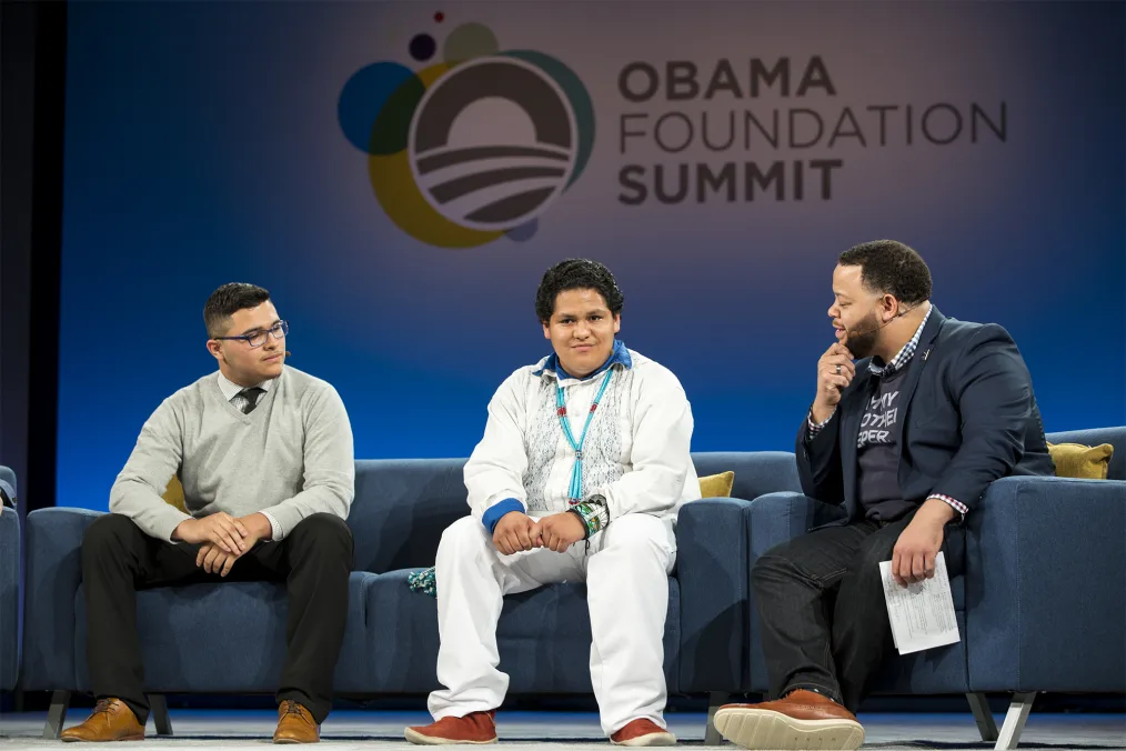 Three men sit on sofas in conversation in front of an Obama Foundation Summit logo