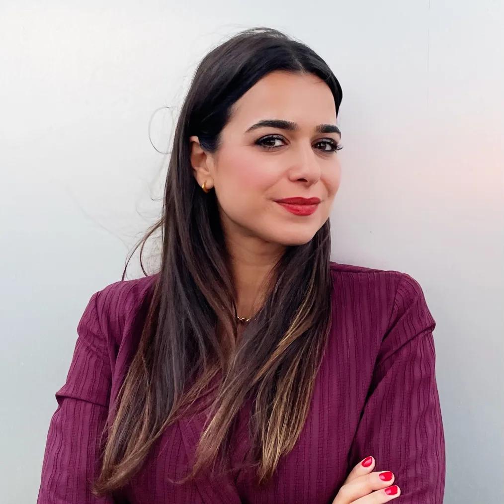 A woman with medium-light skin tone, and long, straight, brown hair poses, unsmiling, with her head turned to the side, staring at the camera. She wears a magenta jacket, bright red lipstick and bright red nail polish. 