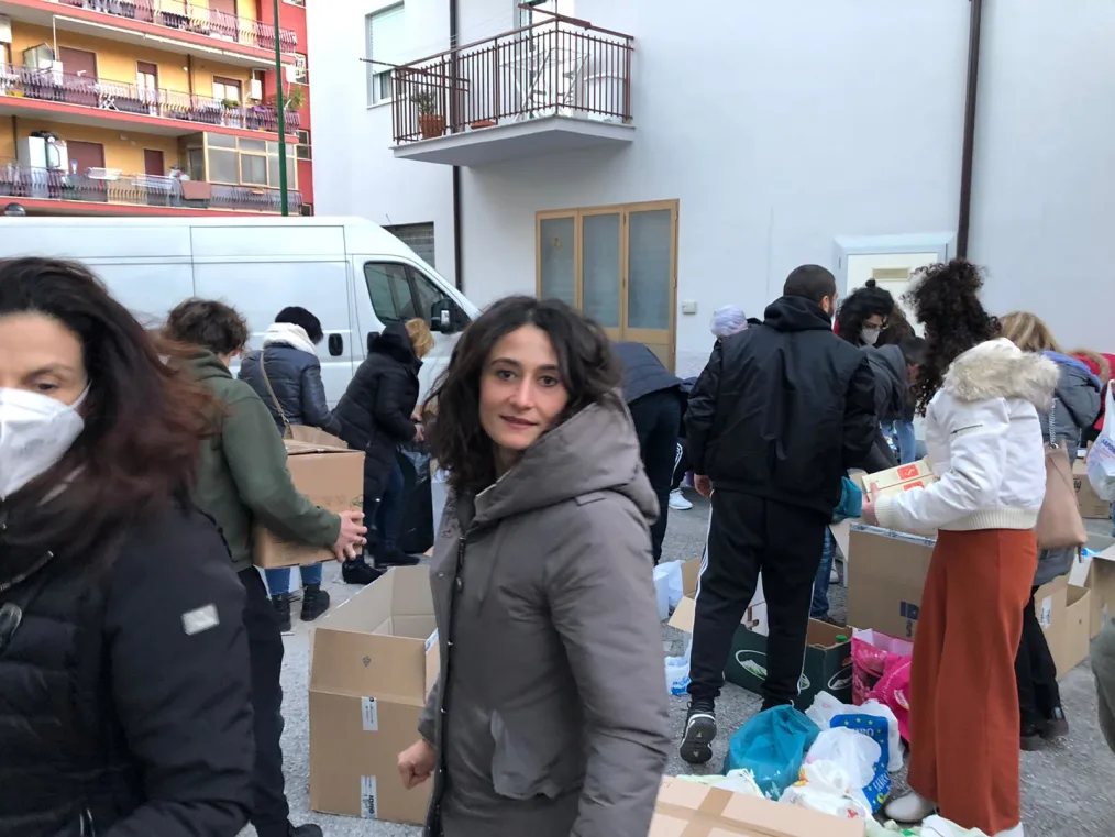 Federica Vinci looks to camera wearing a gray hooded coat. In the background, people are seen standing near boxes and bags of supplies.