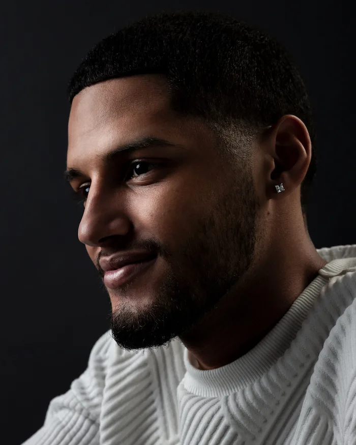 A portrait photo of a man with a light-medium skin tone wearing a white patterned sweater smirking toward the left-hand side of the photo in front of a gradient black background. 