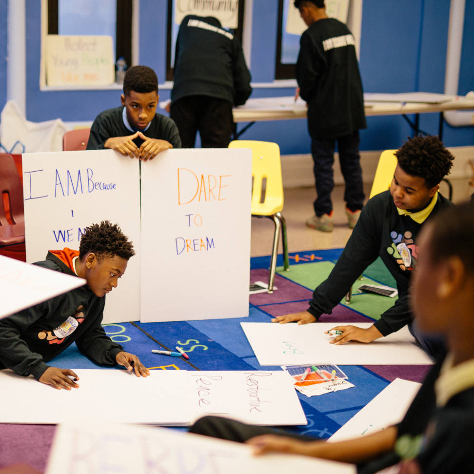 Seven young men in uniforms with a range of light to deep skin tones create posters. The boy in the center has a medium skin tone and is holding two signs that read, “I am because we are'' and “Dare to dream.”