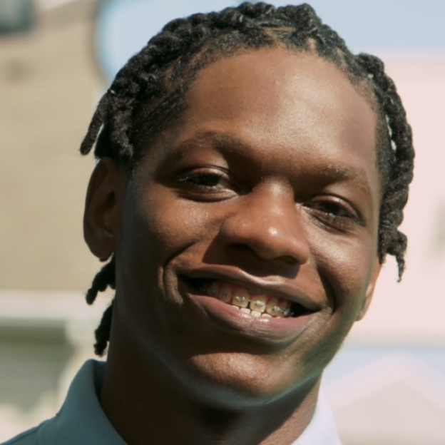 Marquis Wright, a Black boy with a dark skin tone, smiles at the camera. He has braces and locs. 