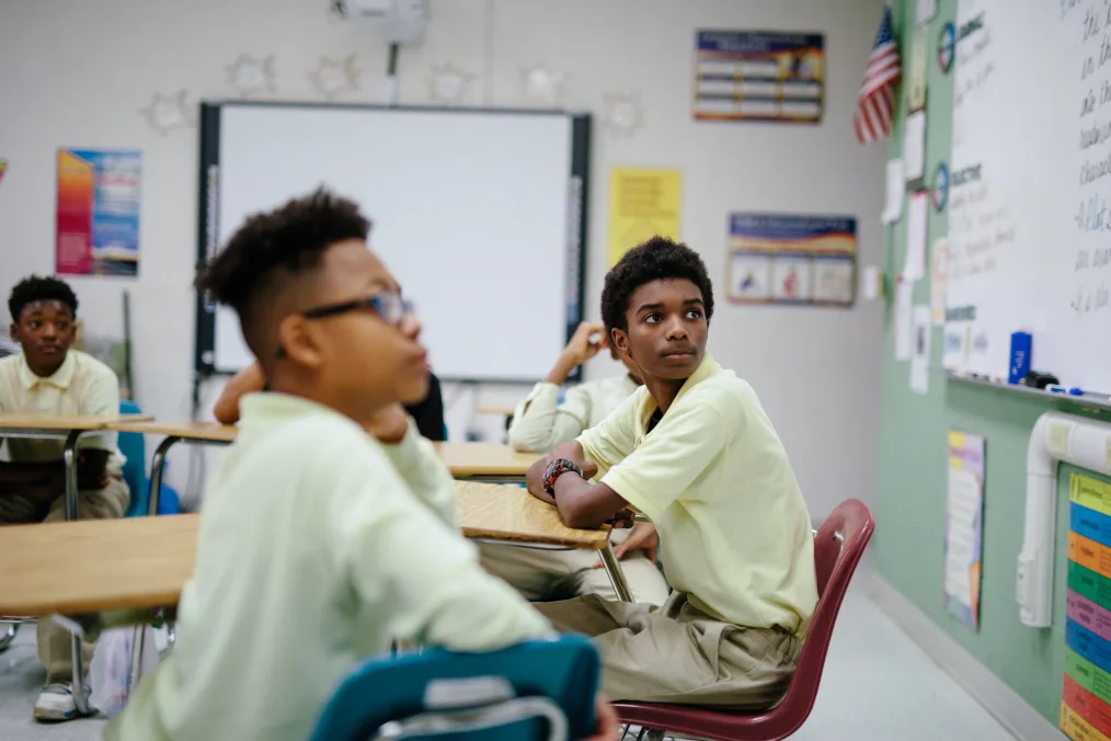 Black boys with a range of light to deep skin tones sit around a table and watch the whiteboard. The students are wearing yellow polos and khaki pants.