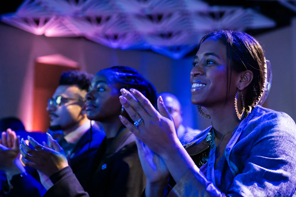 A woman with a dark skin tone claps and smiles in an audience. She is sitting next to a woman with a dark skin tone and a man with a medium skin tone. The room is dimly lit with red and blue lights. 