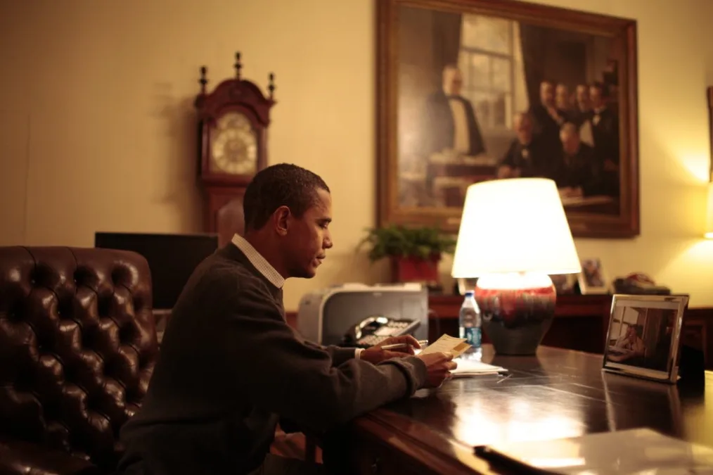 Barack Obama sits in a leather chair at a wood desk in the golden light of a table lamp holding a piece of paper in his hand. Behind him are a grandfather clock and large oil painting.