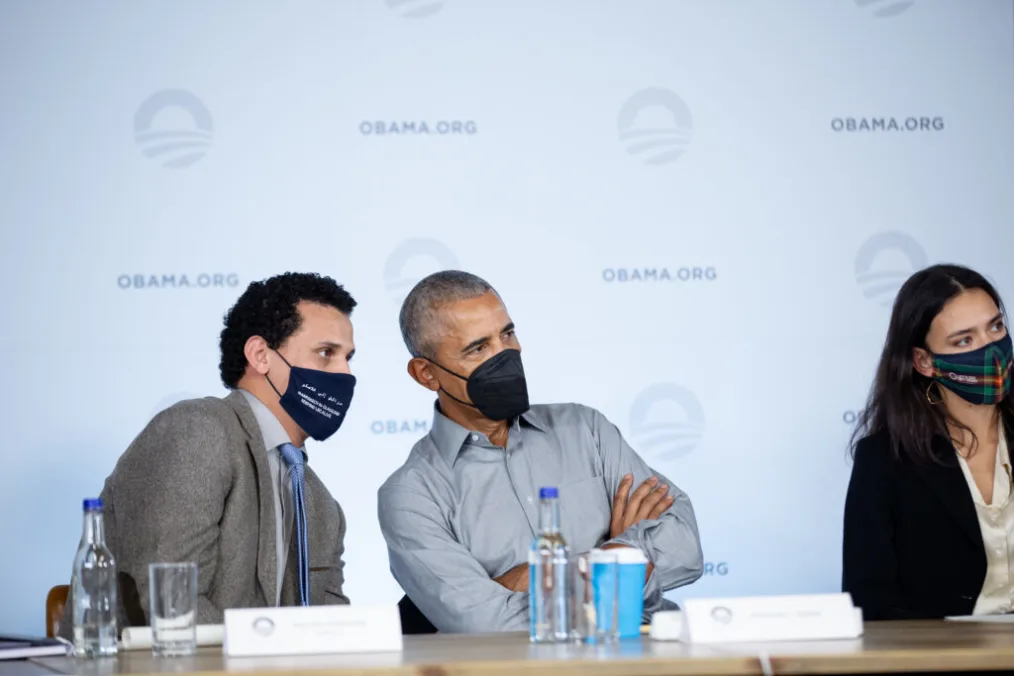 In a gray suit and blue tie and a black mask, Rachid Ennassiri, leans to his left with President Obama nearby, leaning to the right. President Obama wears a black mask and has his arms crossed.