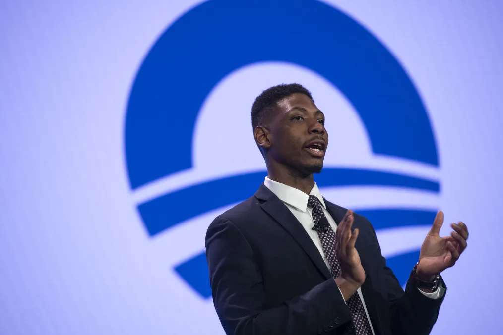 In this portrait, a black man with a deep skin tone and low hair cut wearing a dark blue suit looks off to the right mid clap. The background features the Obama Foundation logo 