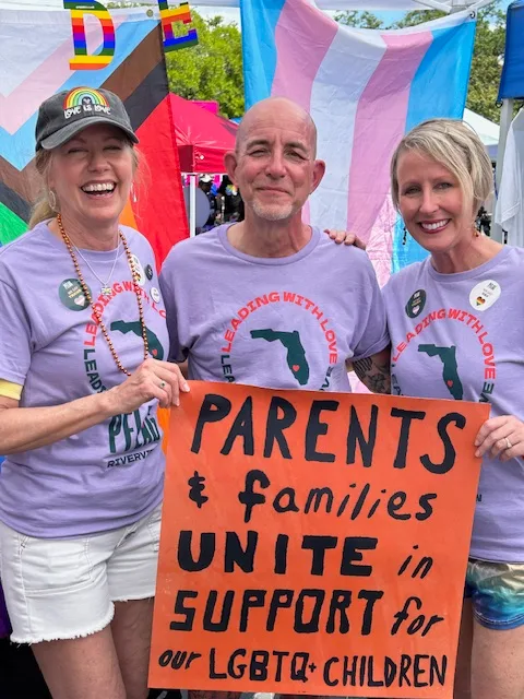 a candid photo of Brian Bond at a PRIDE event. Brian is standing with two other people holding up an orange sign that says, "Parents & families unite in support for our LGBTQ+ children"
