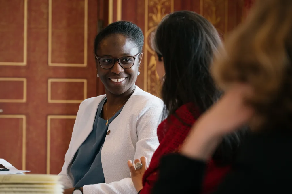 Maïmonatou Mar, a Black woman with a deep skin tone and short black hair, smiles as she speaks to someone. She is wearing glasses and a white blazer. 