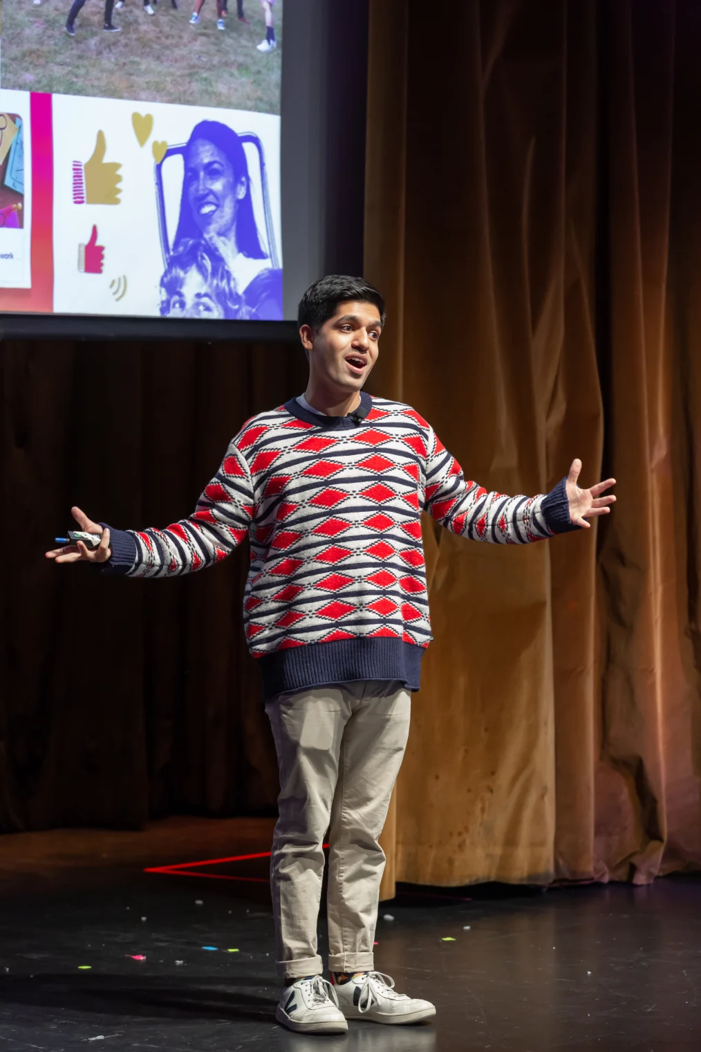 Ziad Ahmed, a man with a medium skin tone, stands on a stage and speaks to an audience. He is wearing a red patterned sweater and his arms are outstretched. A projector screen is behind him. 