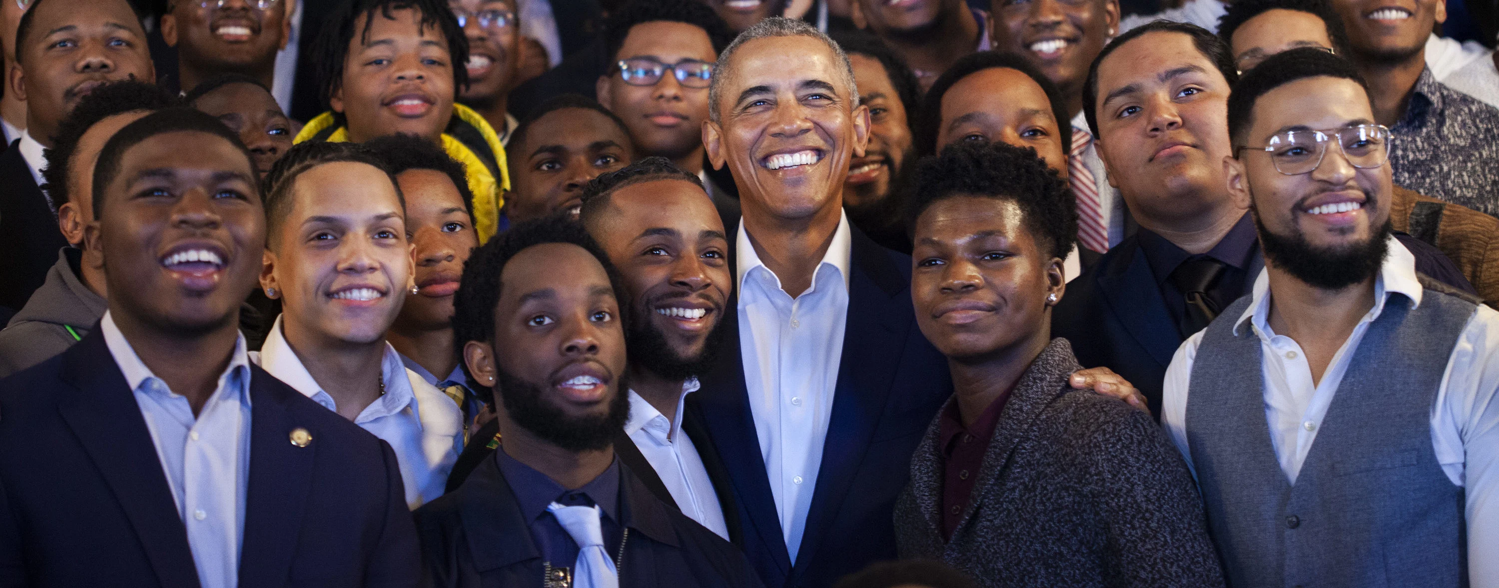 President Barack Obama wearing a white button-up shirt and navy blue blazer smiles amongst a crowd of young men with a variety of skin tones. 