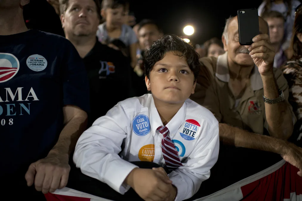 A young boy with a medium skin tone watches President Obama from a crowd. He is wearing a white shirt and red and white striped tie. On his chest are stickers that read, “Cada Voto Cuenta” and “Gotta Vote.” People are gathered around behind him.