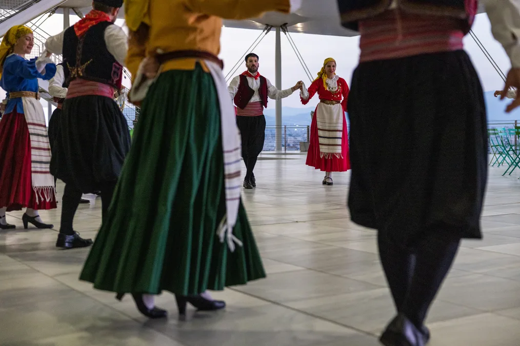A troupe of traditional Greek dancers of varying skin tones are holding hands while dancing. They are on a terrace and wearing bright colors.