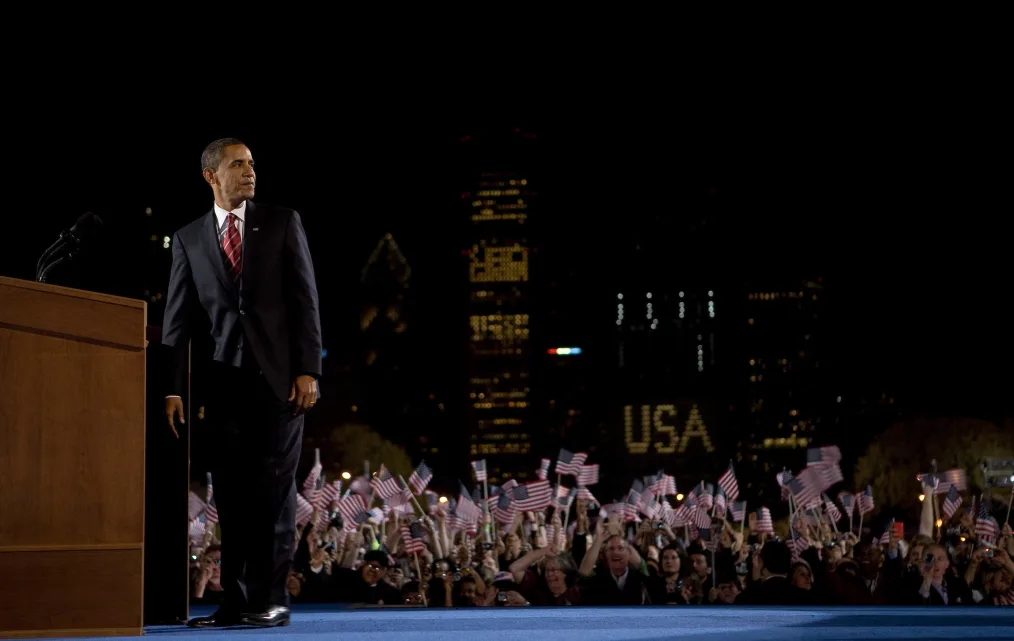 This picture shows President Obama on a big stage wearing a suit and tie while many people behind
him are waving small American flags in the air with tall skyscrapers in the background.